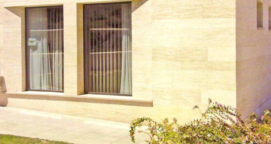 Dos ventanas con persianas verticales de color beige ubicadas en un edificio de piedra de color beige claro con arbustos.