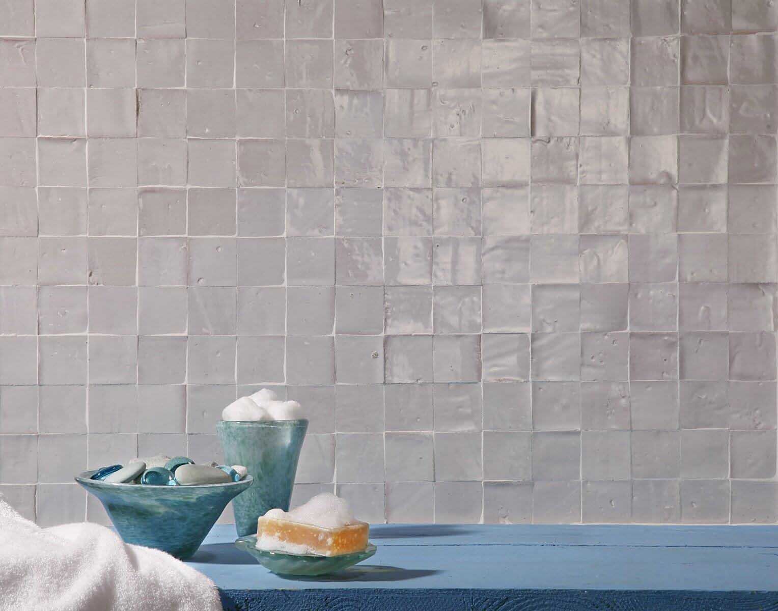 Pared de azulejos cuadrados iridiscentes, cuencos turquesas con jabón y sales de baño sobre una superficie azul.