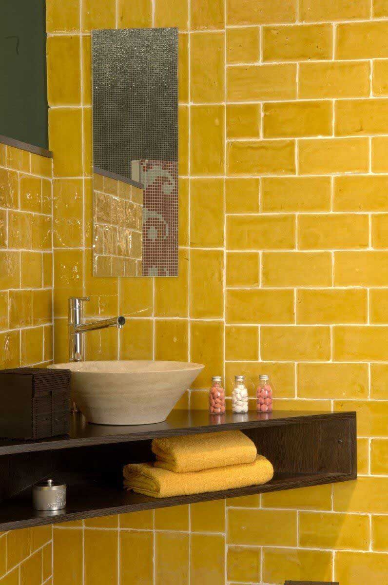 Baño con azulejos amarillos, lavabo, espejo y estante para guardar toallas y artículos de tocador.