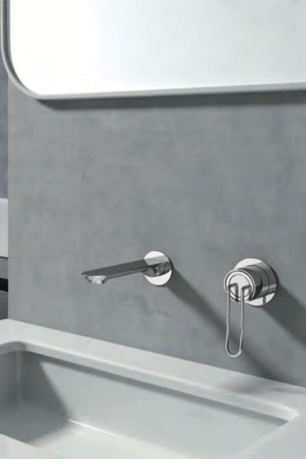 Grifo y manija cromados en una pared de hormigón gris sobre un lavabo blanco y debajo de un espejo.
