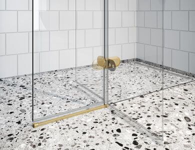 Cabina de ducha con puerta de cristal, herrajes dorados, sobre suelo de baldosas de terrazo, paredes de baldosas blancas.