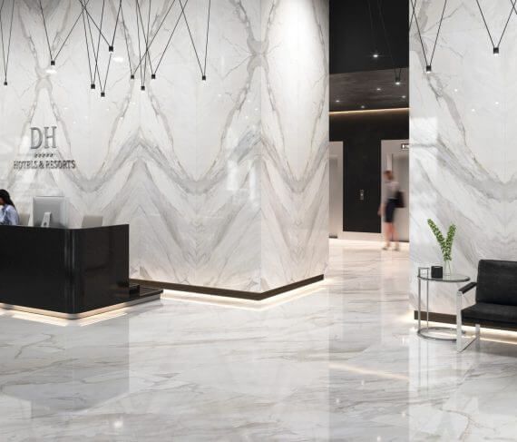 Lobby con paredes y pisos de mármol, mostrador de recepción negro, persona cerca de ascensores, iluminación moderna.