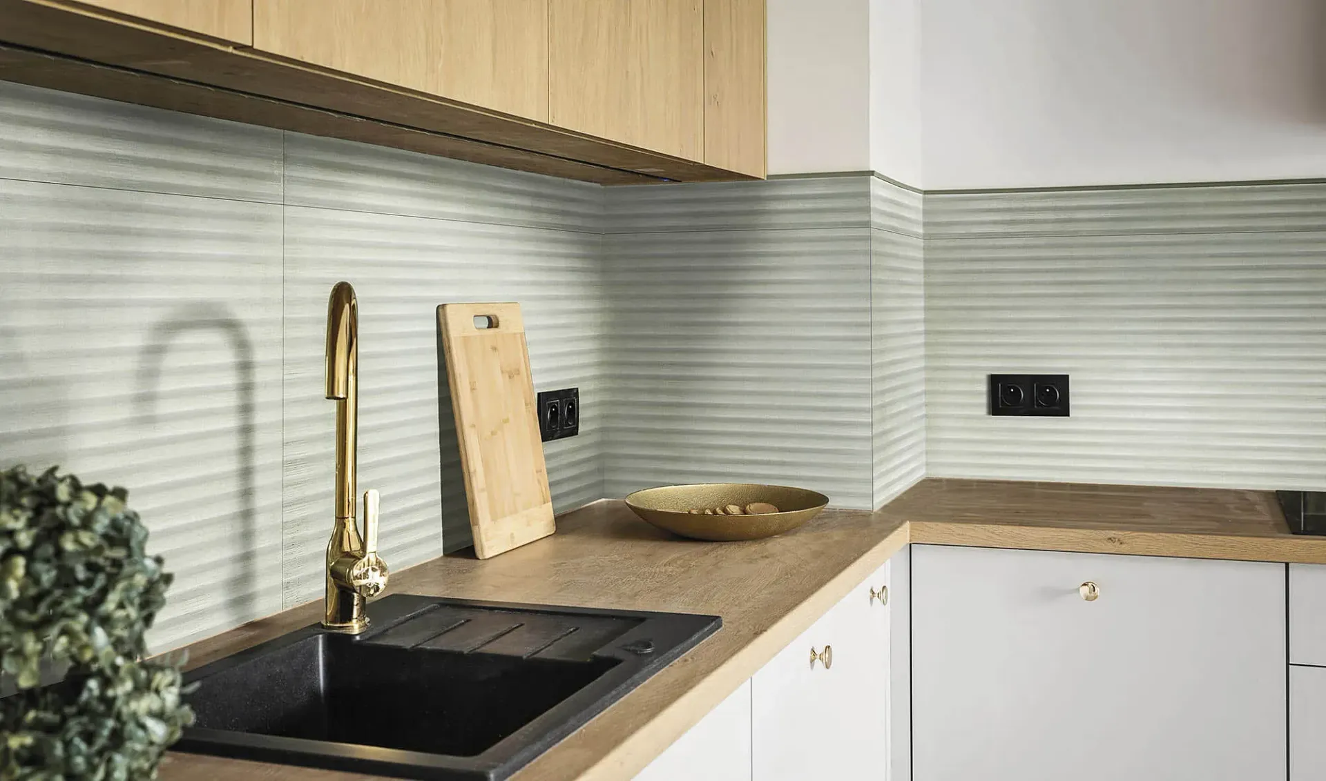 Cocina con grifo dorado, fregadero negro, gabinetes y encimera de madera clara y protector contra salpicaduras texturizado.