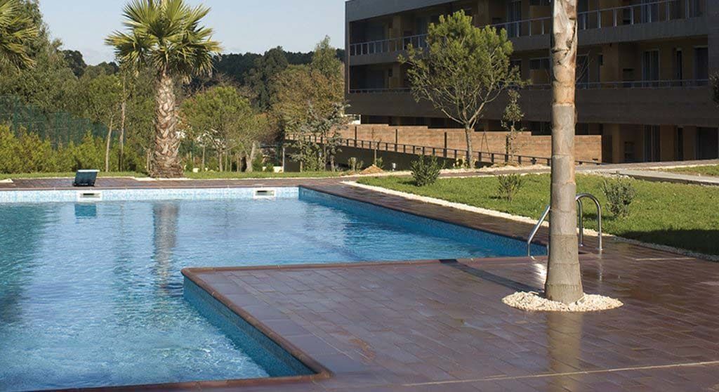 Vista a la piscina, con árboles y el edificio al fondo. Terraza marrón con palmeras.