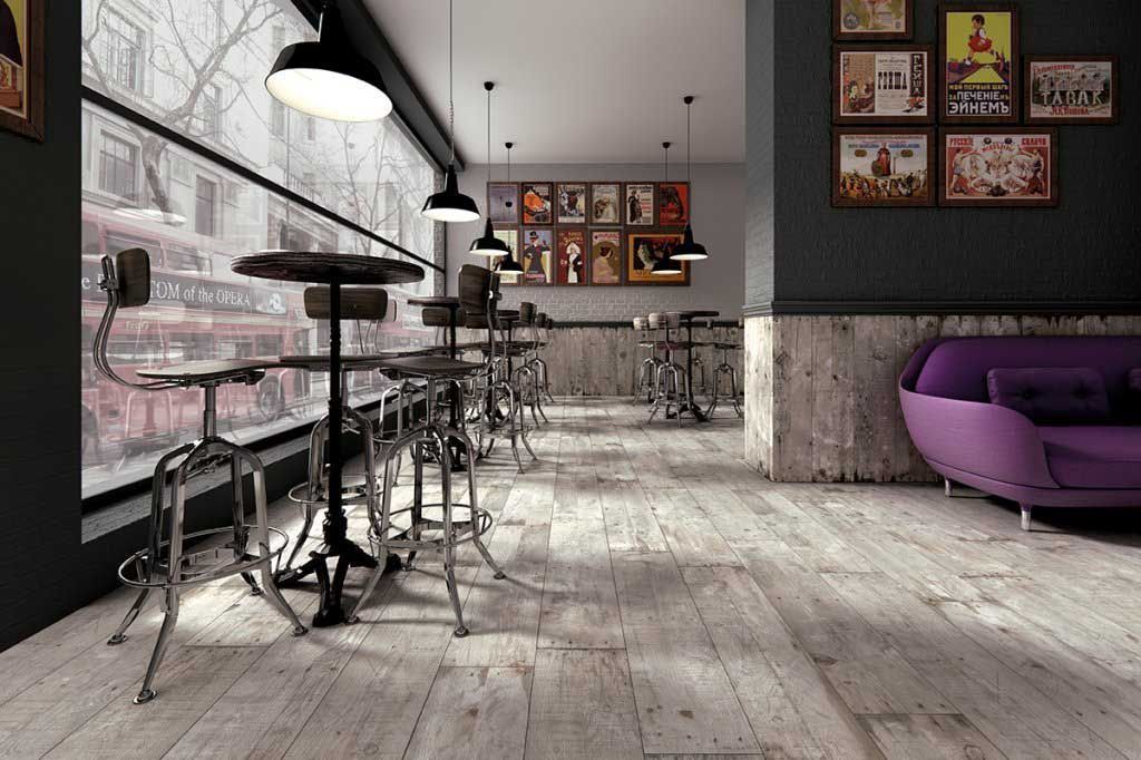 Interior de cafetería con mesas altas, un sofá morado, carteles y una gran ventana con vistas a la calle.