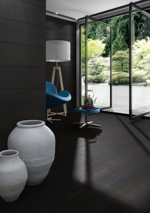 Habitación oscura con puertas abiertas a un jardín, cerámica de color claro, silla azul, piso de madera, lámpara alta.