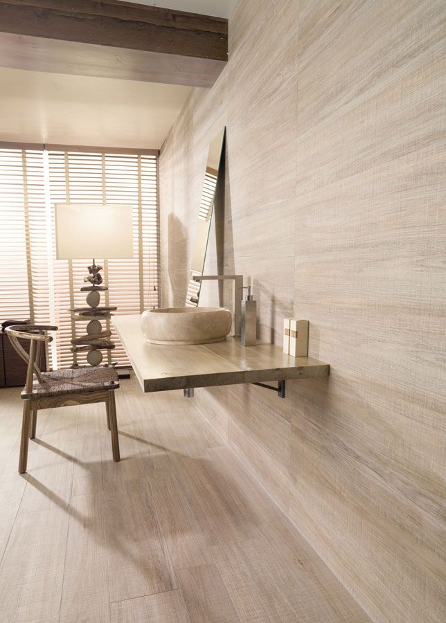 Baño con tonos de madera clara: tocador flotante, espejo grande, silla de madera y pared texturizada.