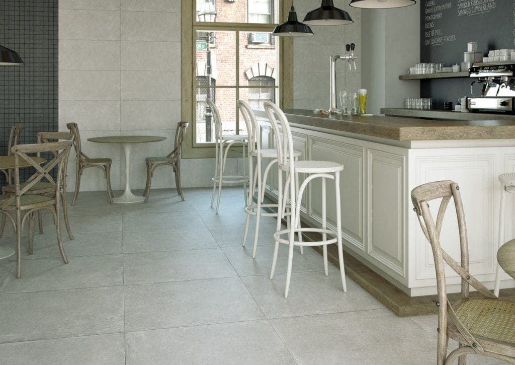 Interior de una cafetería con paredes y mostradores blancos, sillas de madera y una ventana que da a un edificio de ladrillo.
