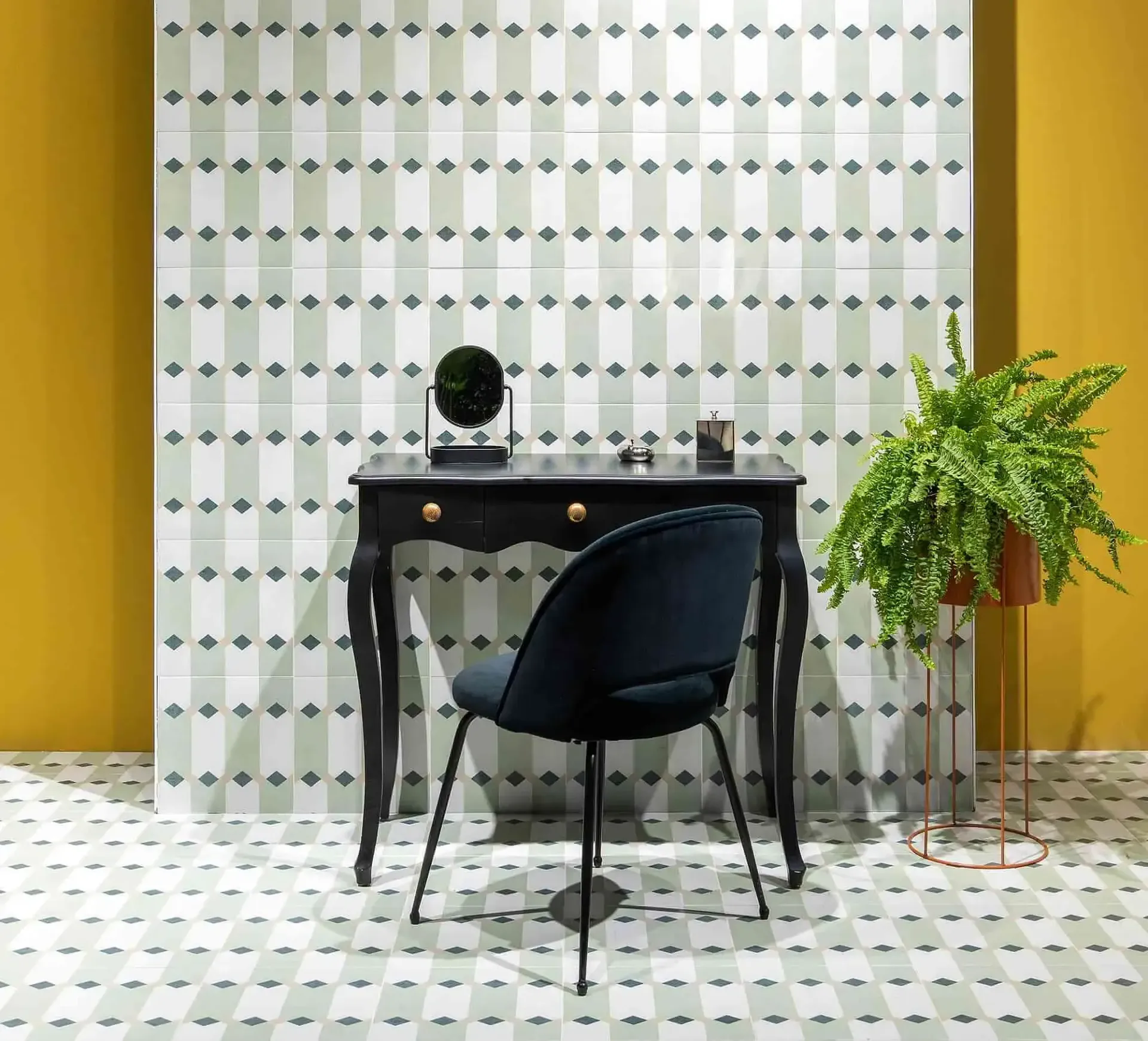 Tocador con mesa y silla negras, pared y piso de azulejos estampados, paredes amarillas, helecho en maceta.