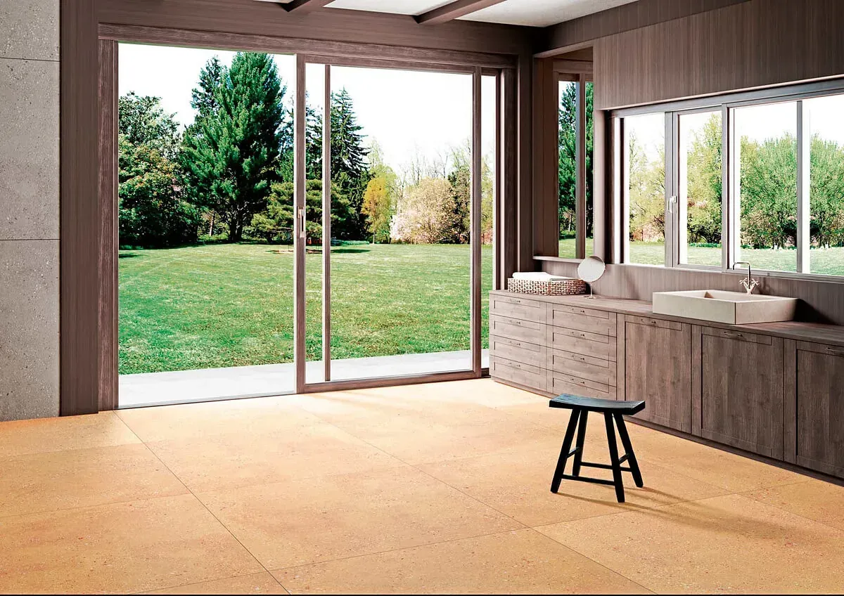 Baño con grandes ventanales con vistas a un césped verde y árboles; paneles de madera, taburete y suelo de baldosas de color neutro.