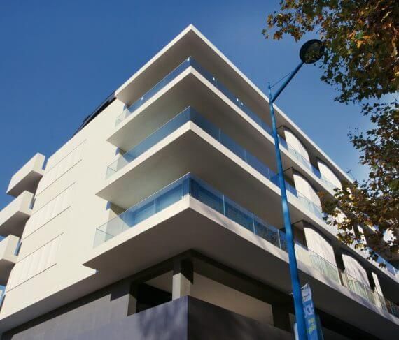 Edificio blanco moderno con balcones salientes y barandillas de cristal azul. Farola y árboles visibles.