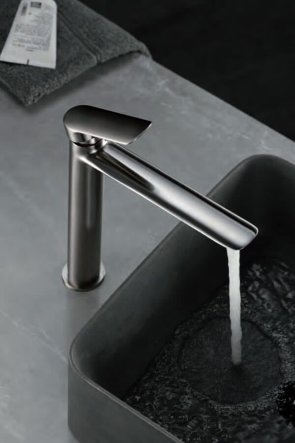 Grifo plateado cepillado que vierte agua en un lavabo cuadrado gris oscuro. Encimera gris y toalla al fondo.