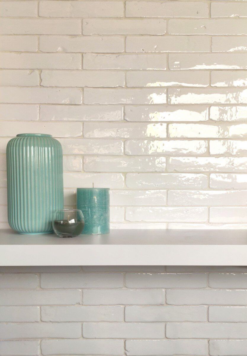 Pared de ladrillo blanco con un estante blanco que sostiene un jarrón verde azulado, una vela y un pequeño recipiente de vidrio.