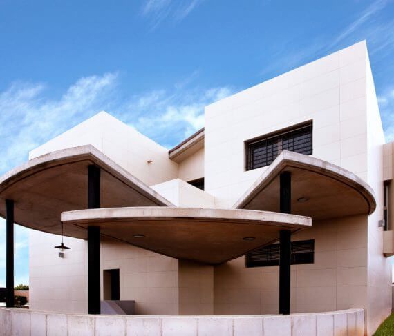 Edificio blanco moderno con aleros de hormigón curvados, columnas negras y pequeñas ventanas, contra un cielo azul.