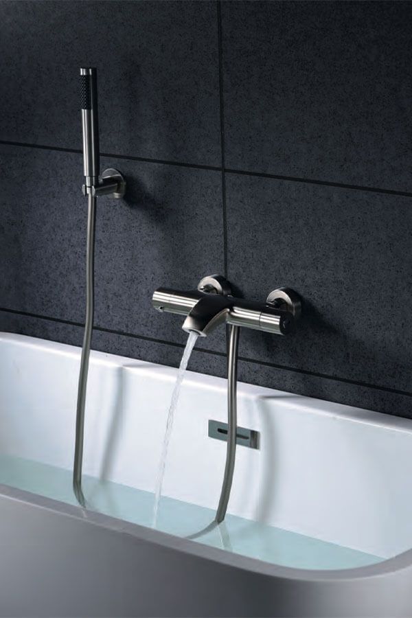 Bañera llenándose de agua desde un grifo cromado y una ducha de mano sobre azulejos oscuros.