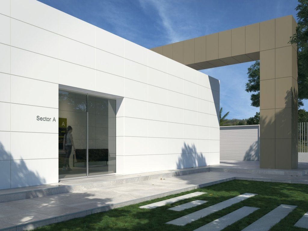 Edificio blanco con una puerta de vidrio, un arco color canela y un camino de concreto que conduce a él, con césped y cielo azul.