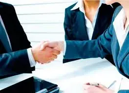 Dos hombres de negocios se dan la mano sobre una mesa; otros están sentados cerca.