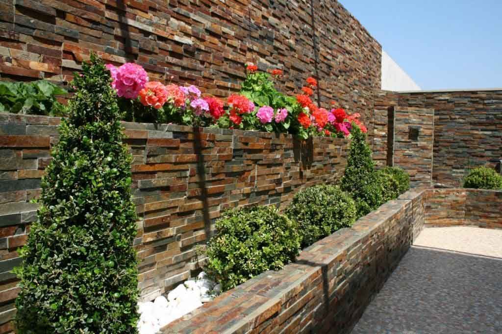 Muro de piedra con jardineras integradas llenas de diversos arbustos verdes y flores de colores.