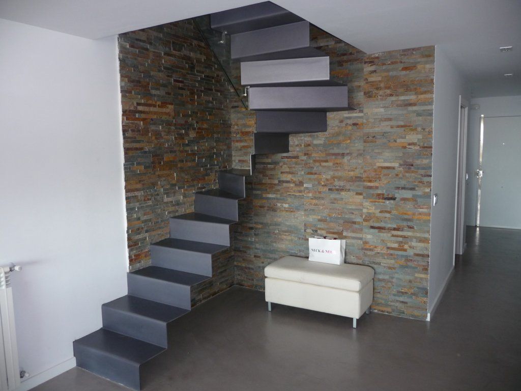 Escalera moderna con escalones grises contra una pared de piedra. Un banco debajo.