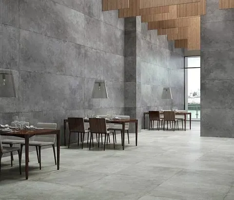 Interior de restaurante con paredes de hormigón gris, mesas, sillas y ventana con vista a una zona verde.