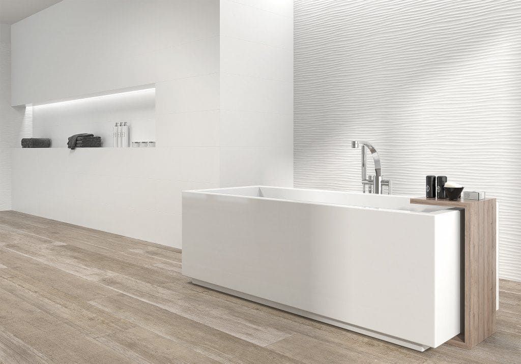 Baño blanco moderno con bañera rectangular, piso de madera y pared texturizada.