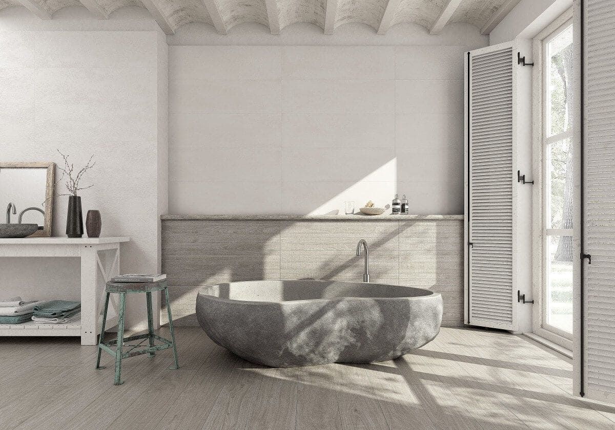 Baño con bañera de piedra, contraventanas de madera y tocador; paleta de colores neutros.