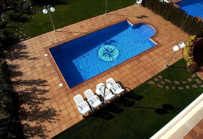 Piscina de agua azul y diseño de brújula, rodeada de azulejos marrones y tumbonas.