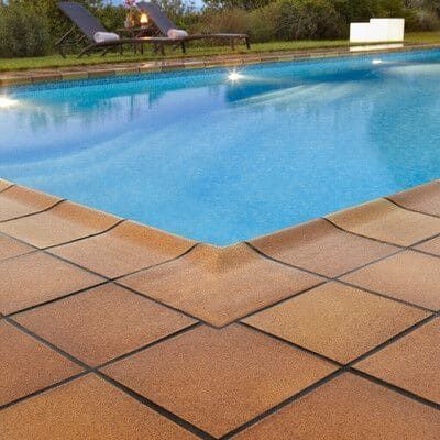 Esquina de una piscina con borde de azulejos color canela; agua azul. Dos tumbonas visibles al fondo.