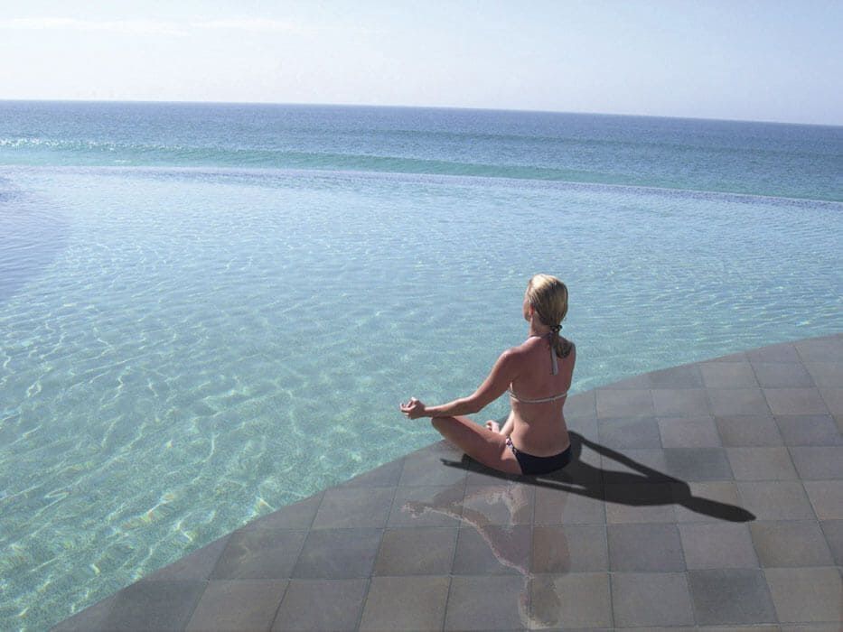 Mujer en bikini meditando junto a la piscina infinita con vistas al océano.