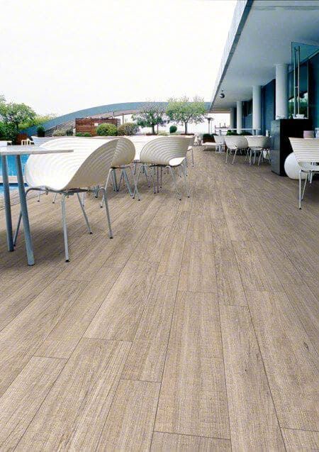 Comedor al aire libre con suelo de imitación madera clara, sillas y mesas blancas. Edificio con detalles azules al fondo.