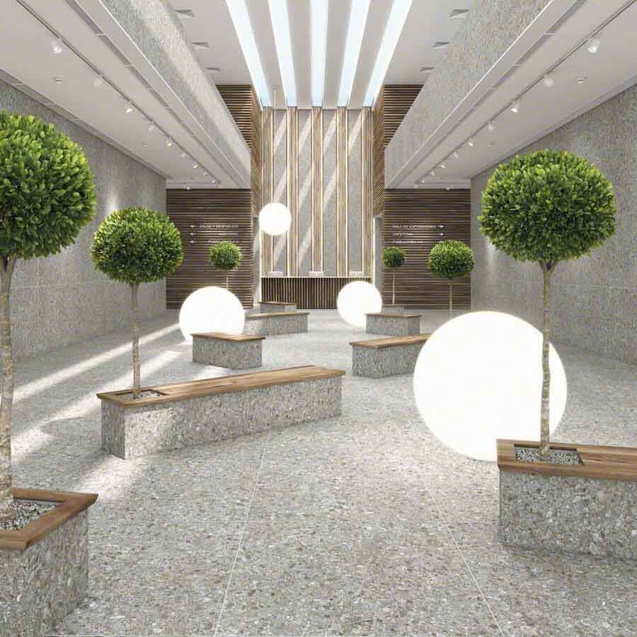 Amplio vestíbulo con suelo de terrazo, bancos, lámparas de globo y árboles topiarios. Tonos neutros.