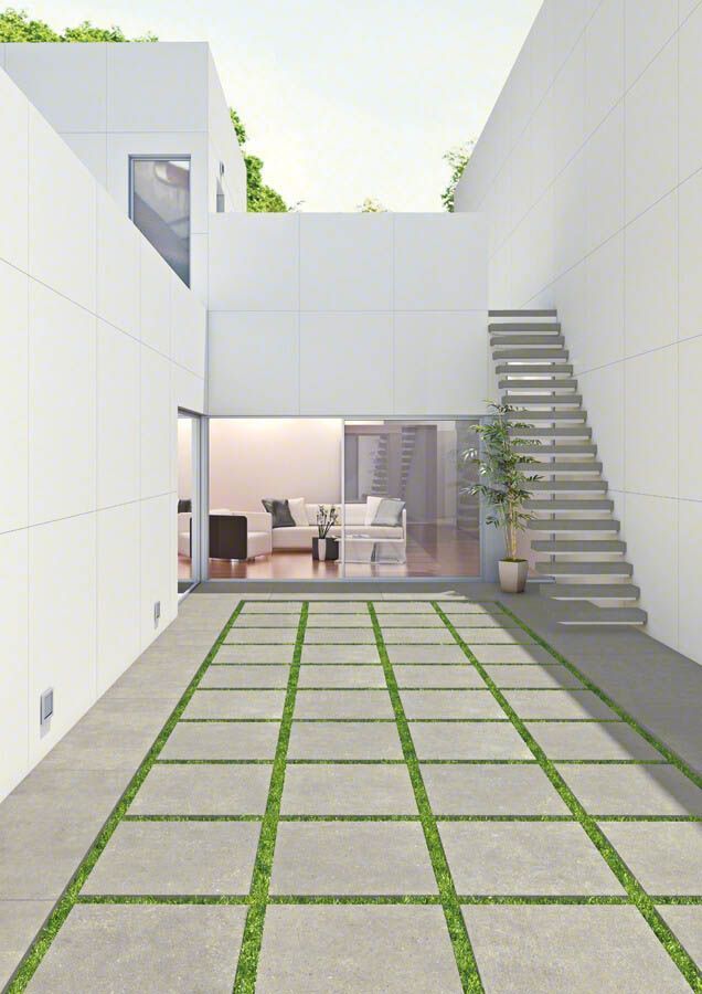 Patio de concreto con inserciones de césped que conduce a una casa blanca moderna, escalera a la derecha, puerta de vidrio abierta al interior.