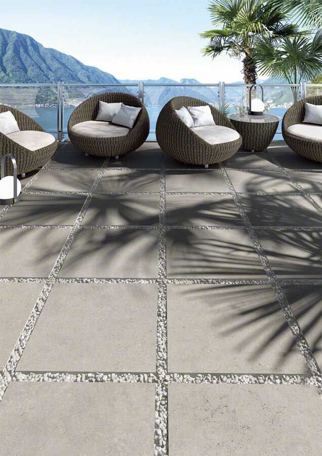 Patio con baldosas de piedra gris, asientos de mimbre y vista al agua. Se ven las sombras de las palmeras.