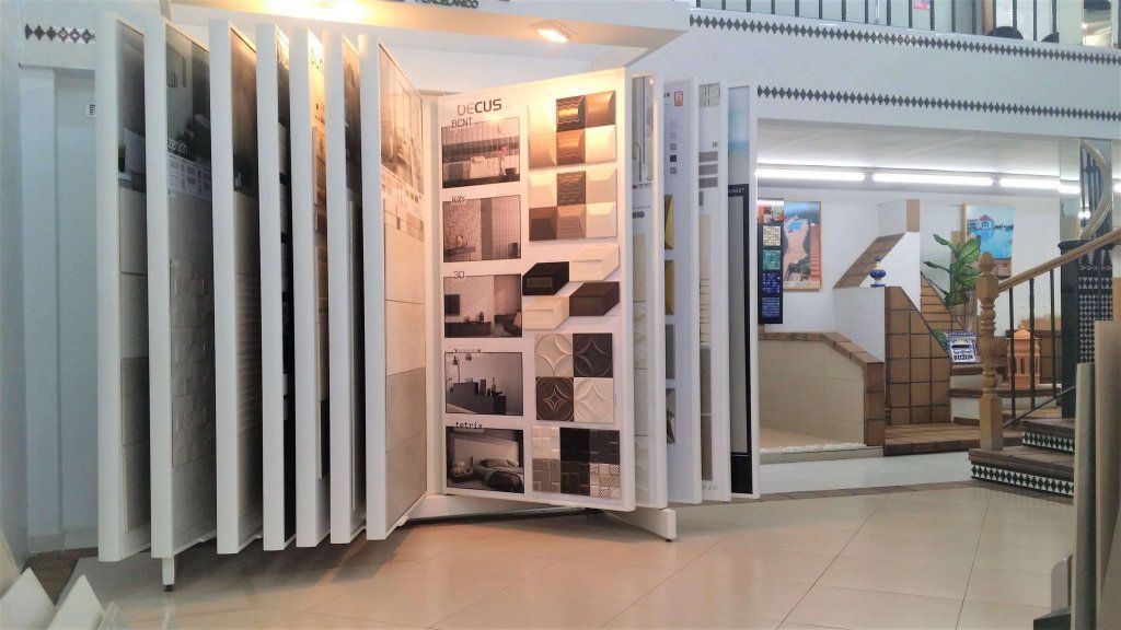 Expositor de azulejos en una sala de exposición, con diversas muestras. Interior luminoso con escaleras y otros expositores.