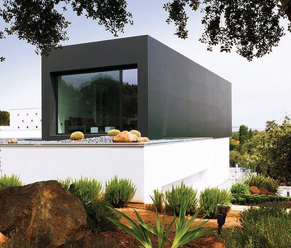 Edificio moderno rectangular de color negro sobre base blanca con un gran ventanal, rodeado de vegetación y rocas.