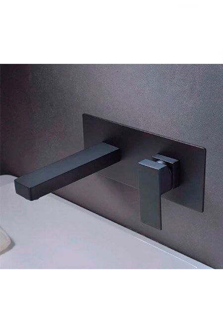 Grifo de pared negro sobre lavabo blanco. Tiene manija plana y caño con placa rectangular sobre pared gris.