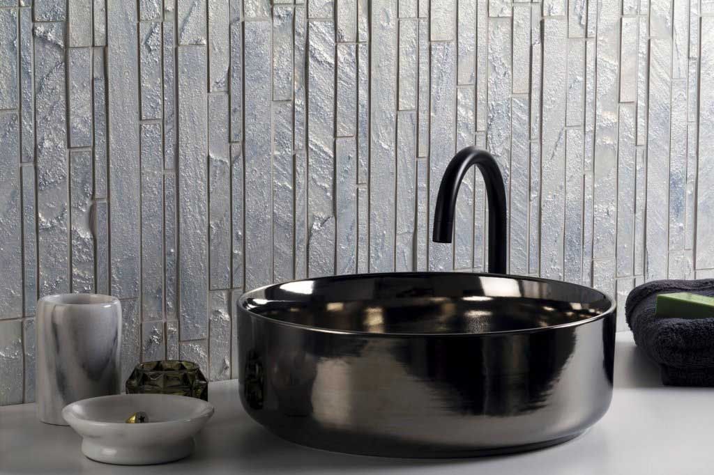 Lavabo de color negro con grifo negro sobre una superficie blanca, contra una pared plateada texturizada.