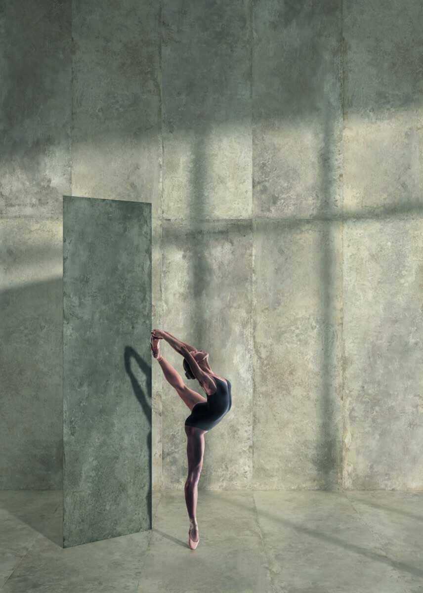 Bailarina en una elegante pose arabesca, en una habitación con una pared de hormigón, proyectando una sombra.