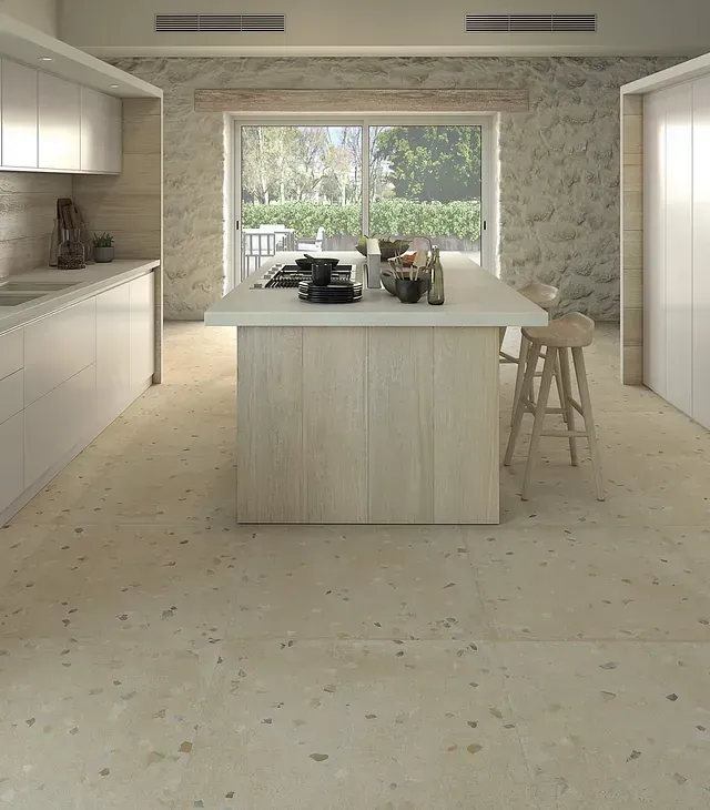 Cocina moderna con isla de madera clara, encimeras y pared imitación piedra. Suelo de terrazo. Gran ventanal con vistas al patio.