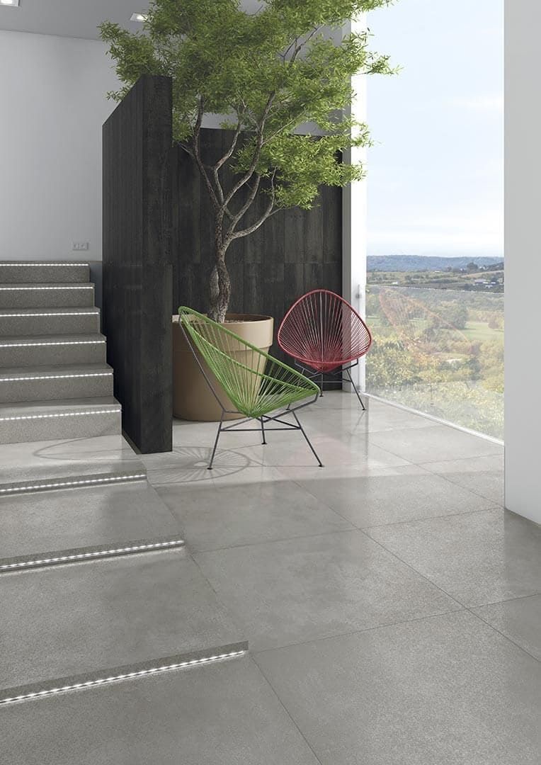 Interior moderno: piso de baldosas grises, escaleras, silla verde, silla roja, árbol en maceta y ventana grande con vista al paisaje.