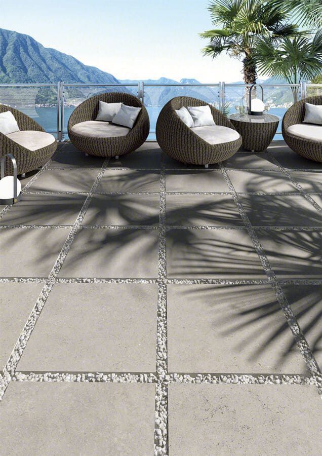 Patio al aire libre con azulejos cuadrados grises, detalles de guijarros, sillas de mimbre y sombras de palmeras.