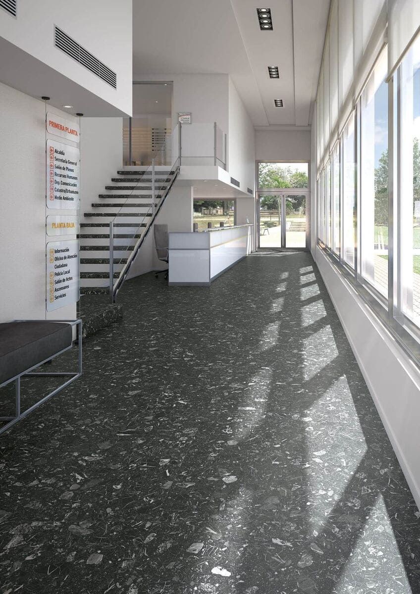 Vestíbulo interior con suelo moteado oscuro, escalera y grandes ventanales con luz solar.