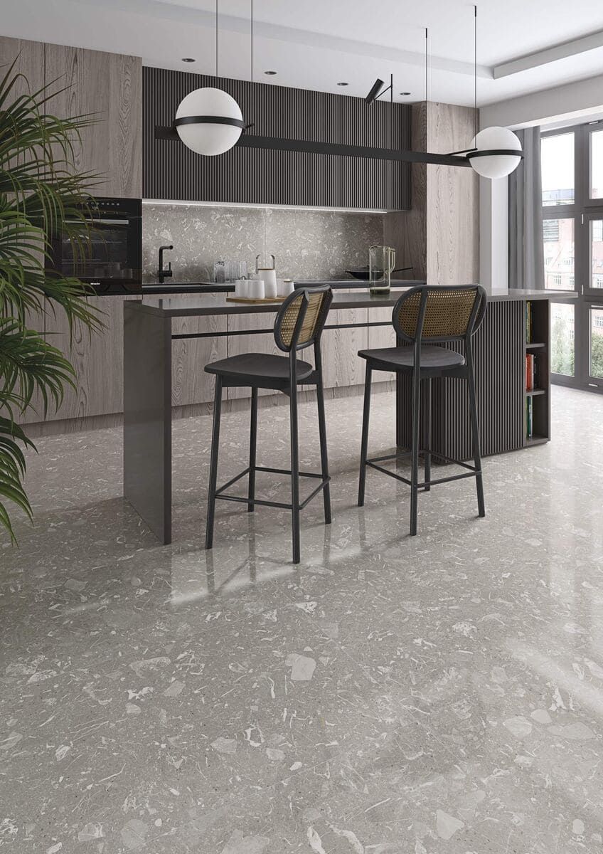 Cocina moderna con suelo moteado gris, isla y taburetes de bar. Armarios de madera y ventana grande.