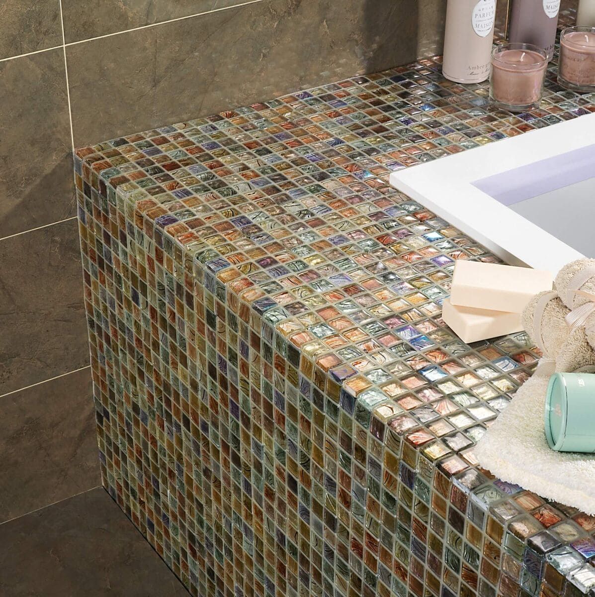 Encimera de baño con revestimiento de mosaico, lavabo blanco incorporado, azulejos de pared marrones y artículos de tocador.