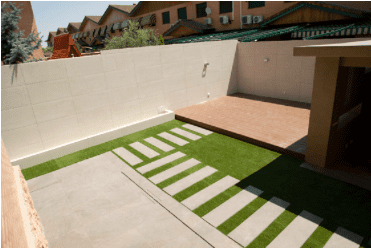 Pequeño patio trasero con césped verde, escalones y una terraza de madera contra una pared blanca.