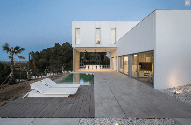 Casa blanca moderna con piscina, patio y tumbonas. Luz nocturna y diseño minimalista.