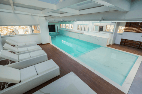 Piscina cubierta con tumbonas blancas, agua azul claro y suelo de madera.