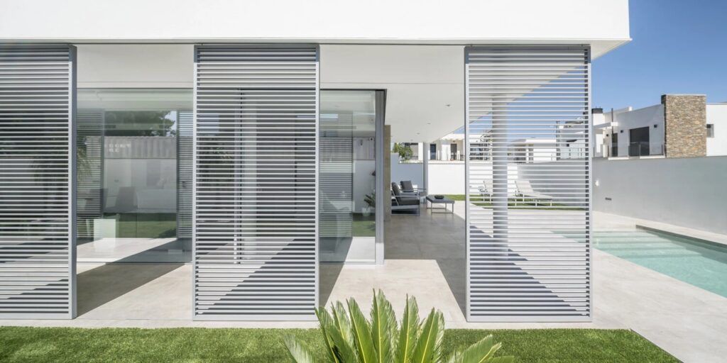 Exterior de casa moderna con paredes de vidrio, persianas horizontales, piscina y césped verde.