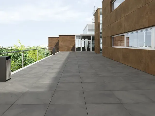 Patio al aire libre con azulejos grises, edificio marrón, barandilla de vidrio y follaje verde.