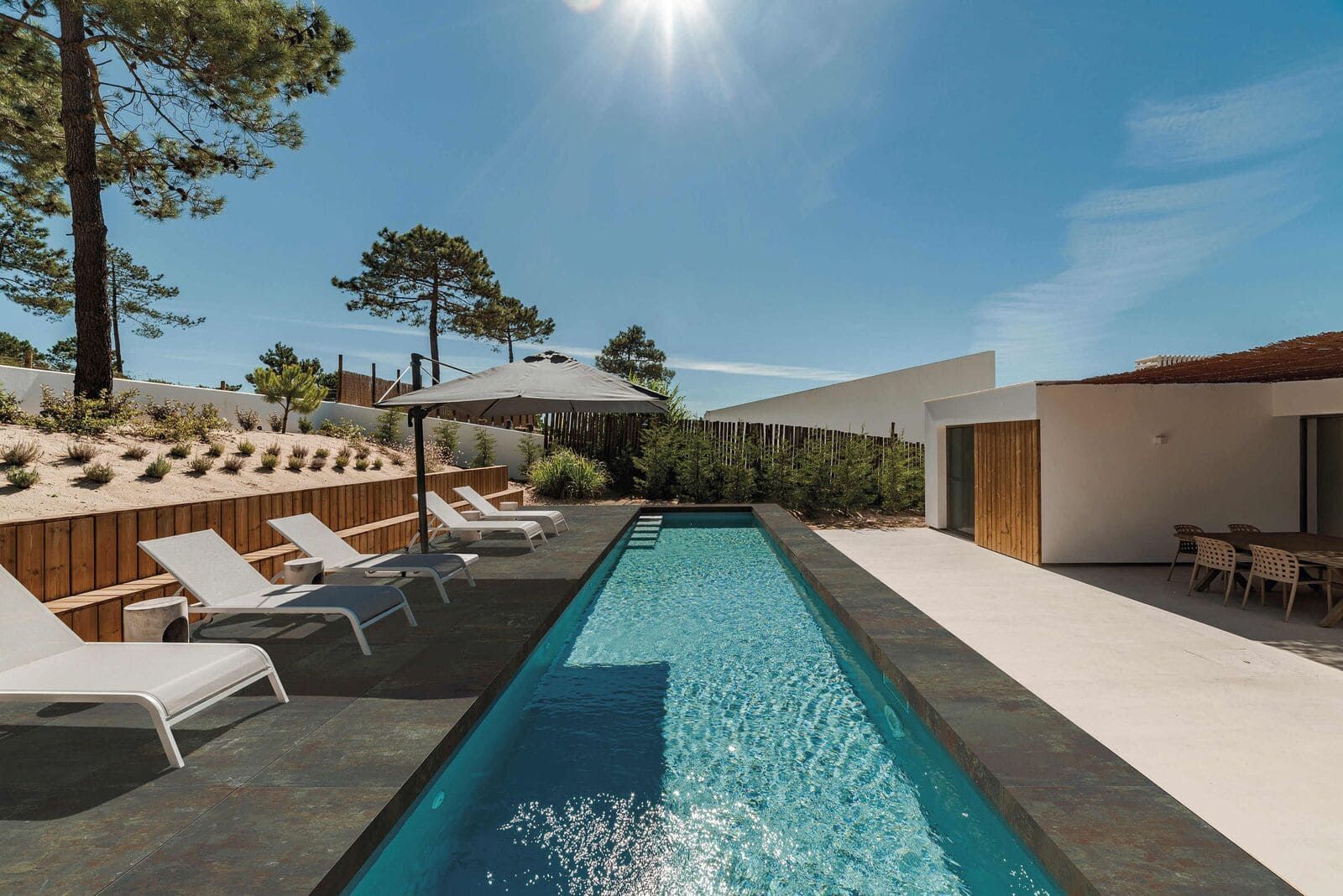 Piscina y tumbonas junto a un moderno edificio blanco bajo un cielo azul soleado.